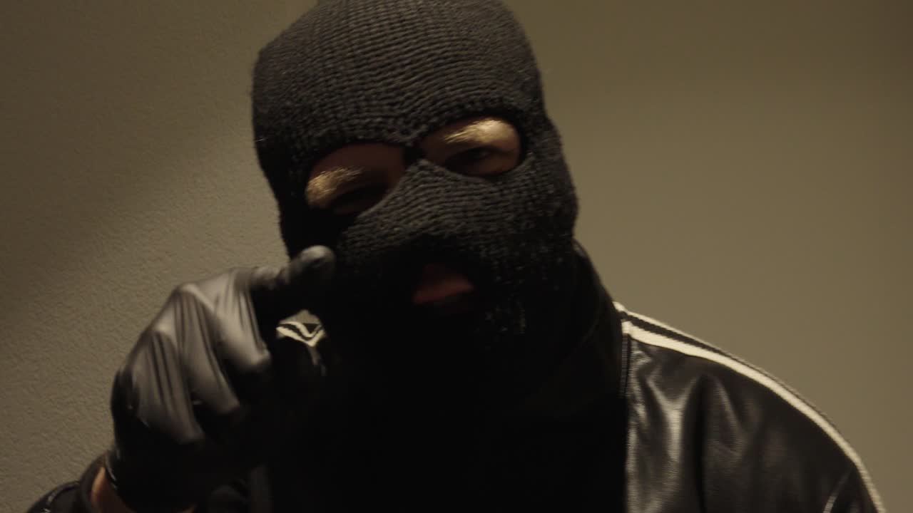 A man wearing a mask and black gloves speaks to the camera, pointing his finger. He threatens and shouts.