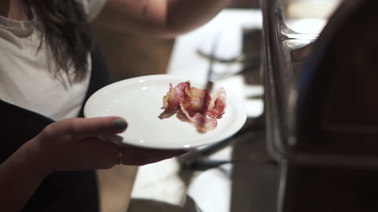 Interesting closeup detailed shot of person serving bacon on plate at hotel breakfast, with stainless steel silverware and full white plate