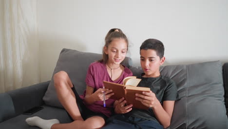 friends sitting comfortably on couch enjoy fun study moment as girl flips book page with cheerful smile while boy reacts playfully making funny face showing joy