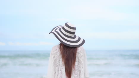 Vista-Trasera-De-Una-Mujer-Con-El-Pelo-Largo-Y-Un-Sombrero-Flexible-Frente-A-Las-Olas-Del-Mar-Y-El-Horizonte,-A-Cámara-Lenta