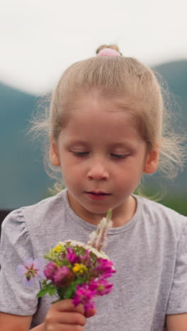 thoughtful little girl with allergy looks at small bouquet of windflowers and sneezes near wooden fence against mountain silhouette closeup