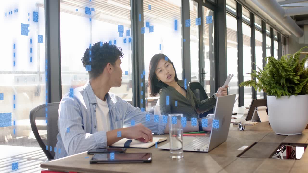 Man writing and glancing at woman's laptop, reaching papers; woman checking docs blue grid aiding