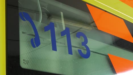 the exterior of yellow paramedic ambulance vehicle, emergency medical service phone number 113 on the glass, medium closeup shot