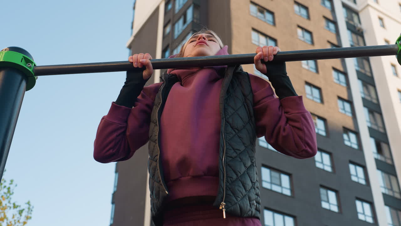 Sky view female exerciser working out intensively on pull up bar against urban building backdrop emphasizing forearm strength focus and control during outdoor calisthenics session