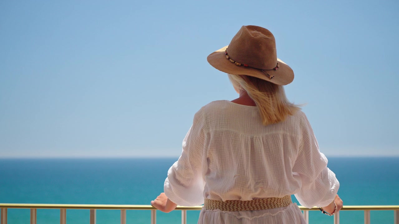 Woman in white dress and straw hat enjoying peaceful sea view from balcony during sunny summer beach vacation.