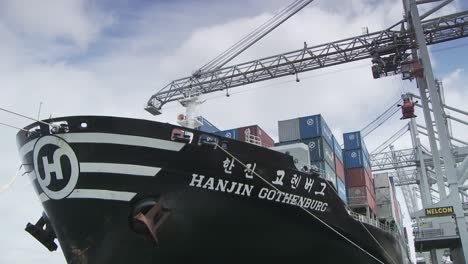 large black cargo ship heavily laden with colorful shipping containers, docked in port under a blue sky with cranes above