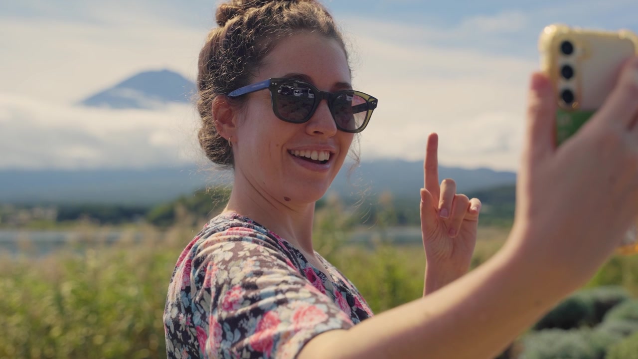 Donna sorridente con occhiali da sole che tiene in mano lo smartphone, registra un video di sé stessa o si scatta un selfie con l'iconico Monte Fuji alle sue spalle, salutando e gesticolando felicemente verso la fotocamera.
