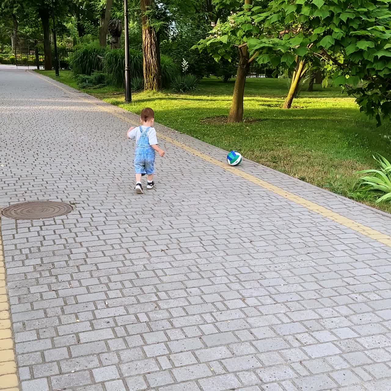 Baby boy goes to pick up the ball from the grass. Kid brings his ball to the camera. Toddler playing in the park