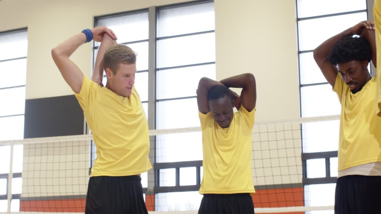 Diverse male teammates in yellow stretching triceps, swinging arms on gym court by net before drill