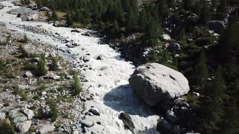 Mountain-muddy-stream-with-a-big-rock-on-the-path,-valley-with-water-from-the-glacier-and-fir-forest,-drone-aerial-view