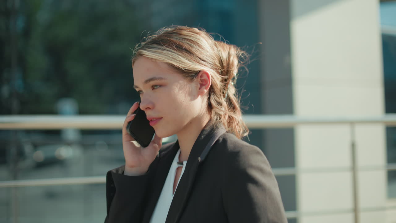 Side view of calm lady in formal outfit talking on phone with eyes closed, walking toward meeting, serene expression, modern glass building background