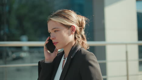 side view of calm lady in formal outfit talking on phone with eyes closed, walking toward meeting, serene expression, modern glass building background