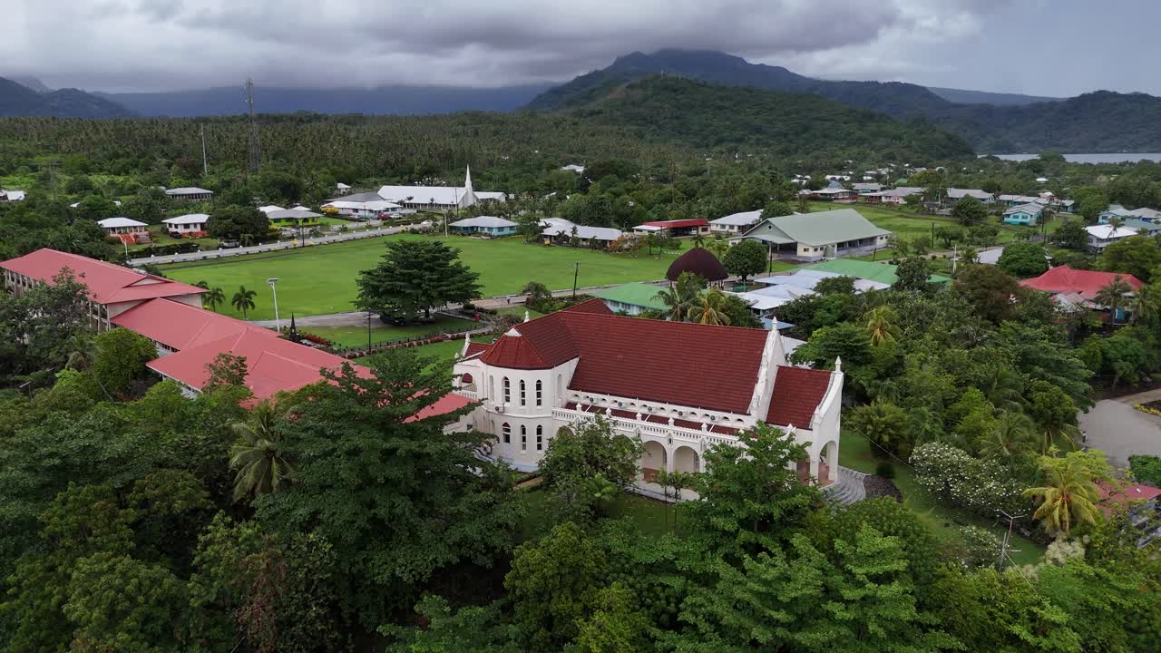 Dronebillede af en traditionel, hvid katedral i en frodig, tropisk landsby i Samoa. Et smukt vartegn omgivet af livlige, grønne Stillehavslandskaber og lokale huse.