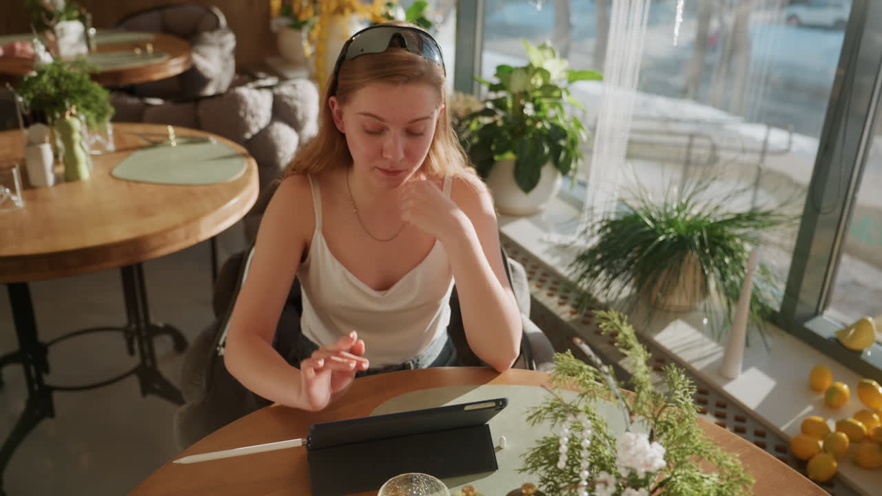 Bílá žena studující na tabletu v prosvětlené kavárně, v ležérním tílku, sluneční brýle na hlavě, rostliny a květinová dekorace na stole, citrony u okna, ranní světlo proudící sklem, soustředěný výraz