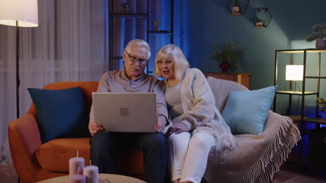 happy senior elderly couple using laptop pc talking together doing online shopping at home sofa