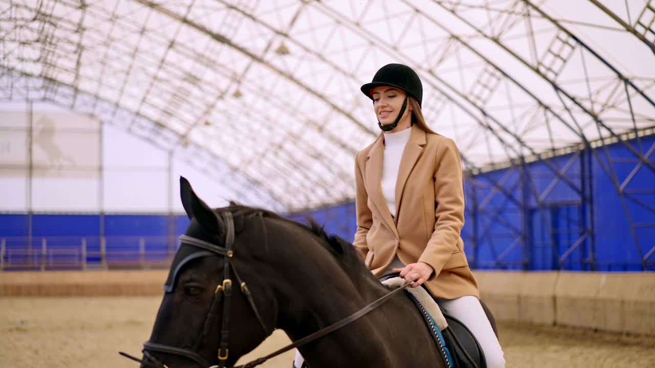 Caucasian woman in black jockey hat practicing horse riding. Lady takes riding lessons on a beautiful black horse. Manege at backdrop in blur.
