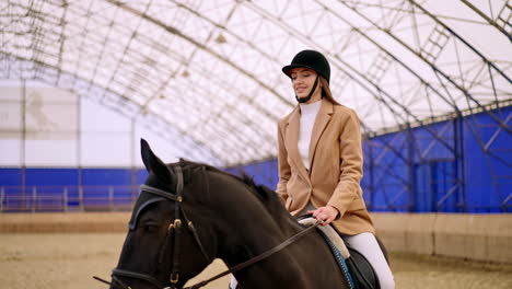 caucasian woman in black jockey hat practicing horse riding. lady takes riding lessons on a beautiful black horse. manege at backdrop in blur.