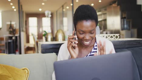 Happy-african-american-woman-sitting-on-sofa-in-living-room,-using-laptop-and-talking-on-smartphone