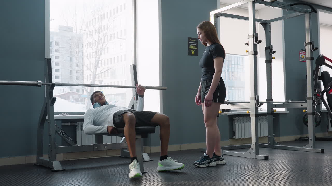 young sister watches brother bench press weighted barbell on flat bench inside modern gym with squat rack free weights and city view through window while coach and bystander exercise in background