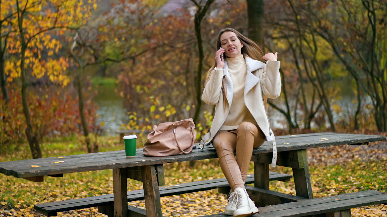 Modern smartly dressed lady sitting on the table outdoors talking on the phone. Beautiful autumn nature and river at backdrop in blur.
