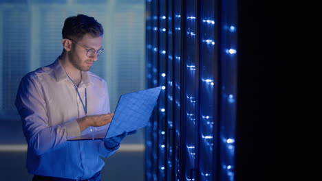 Ingeniero-De-Servidores-Masculino-En-El-Centro-De-Datos.-Ingeniero-De-TI-Inspeccionando-Un-Gabinete-De-Servidor-Seguro-Utilizando-Tecnología-Moderna-De-Coworking-De-Computadoras-Portátiles-En-El-Centro-De-Datos.