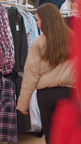una chica con una chaqueta marrón y pantalones negros se ve caminando por una tienda de ropa, llevando una bolsa de gimnasia. captura una vista trasera de ella mientras se mueve más allá de los estantes llenos de varias camisas y pantalóns