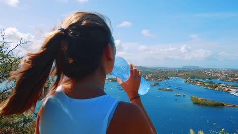 Girl-hiker-drinking-water-bottle-while-admiring-a-harbor-in-Curacao,-Caribbean