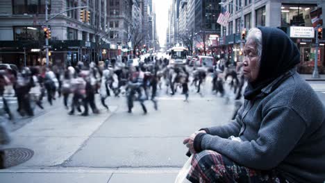 an elderly woman sits alone, wrapped in a shawl, observing the bustling crowd of pedestrians crossing the street in a vibrant urban setting. busy city life unfolds around her as she watches.