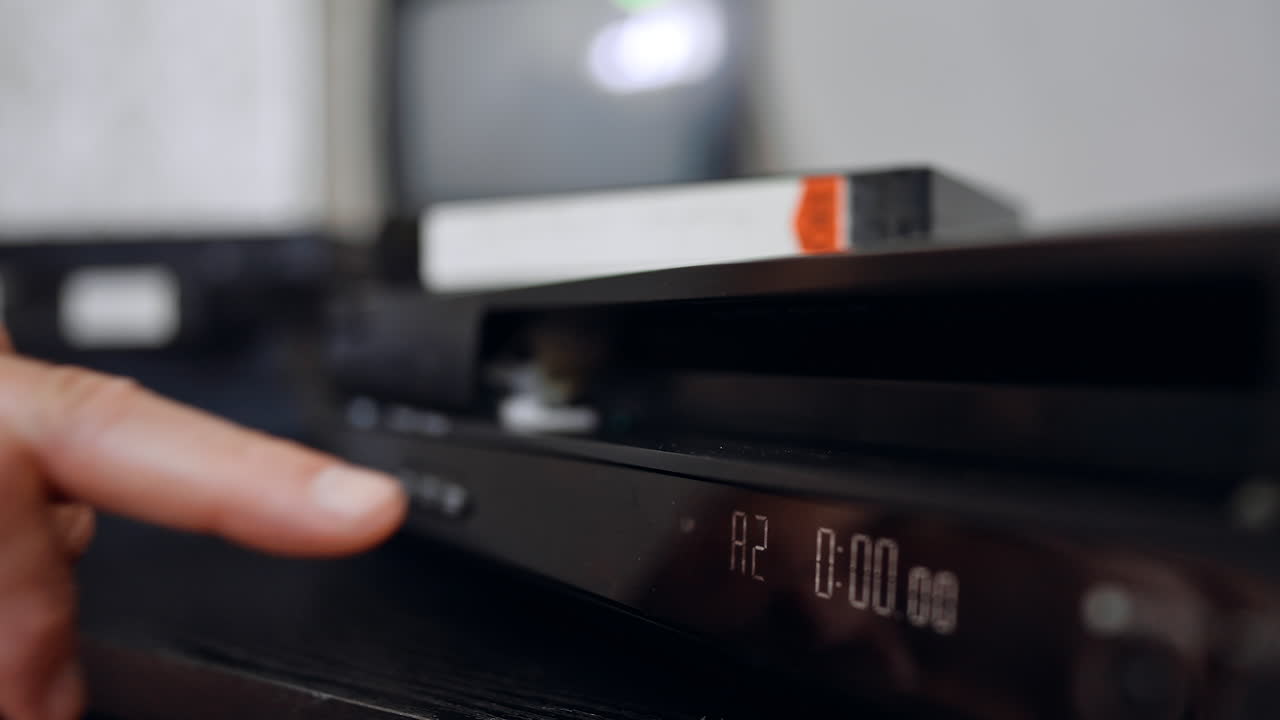 Male hand places the VHS tape into the recorder. Extreme close-up. Screen at blurred backdrop switches on