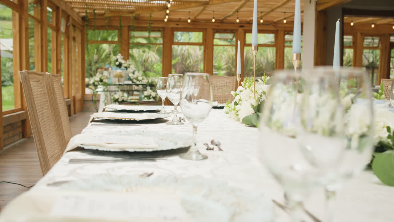 Clip of a finely decorated wedding table with a white tablecloth, glass stemware, shiny cutlery, plates, and a delicate floral arrangement in the center.