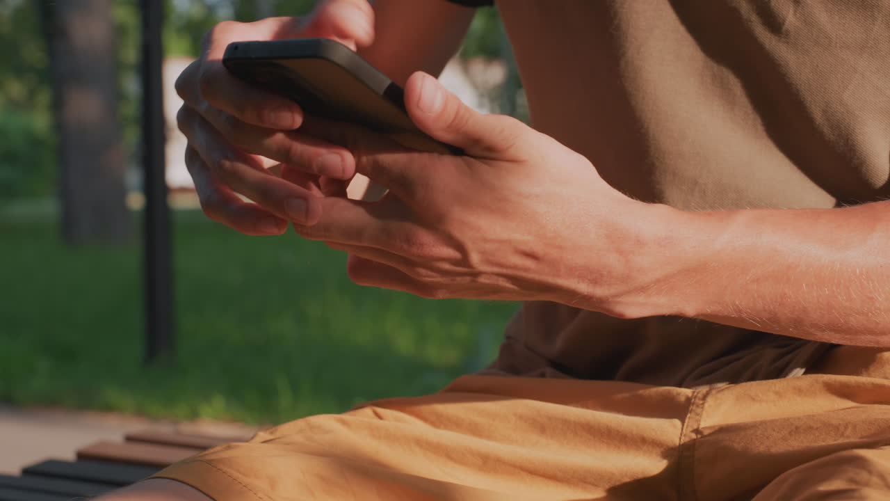 Man On Bench Typing And Scrolling On Phone, Focused Thumbs Composing Content In Warm Daylight, Casual Shorts And Grass Backdrop Emphasize Creative Microinteractions And Social Routine