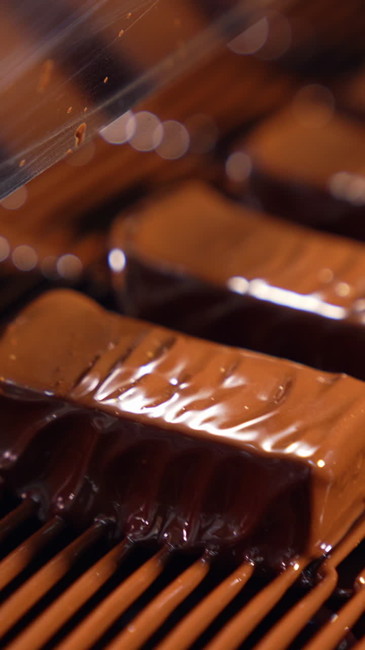 Sweets are poured with chocolate and blown by the airflow. Little waves of chocolate roll along the candies close up. The production line at a candy factory. Vertical video