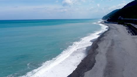 a serene coastline with turquoise waters, gray sand, and a train track running alongside, aerial view