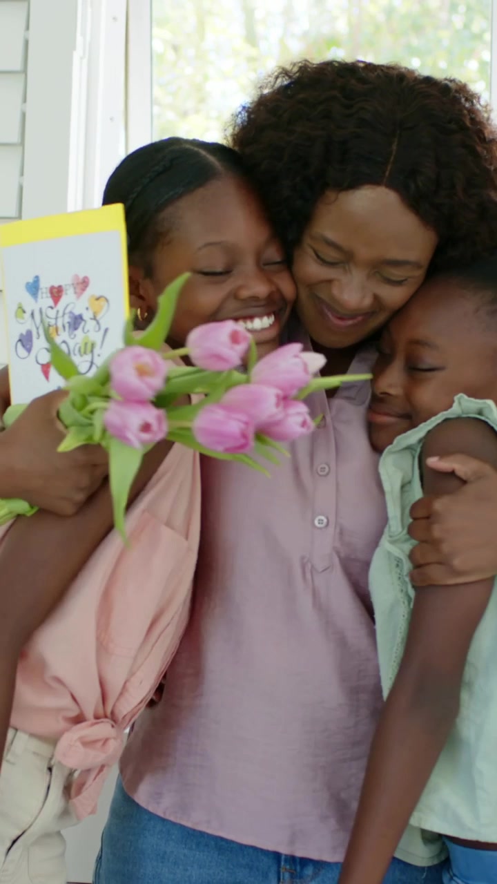 Vídeo vertical: Mãe sorridente de rosa abraçando as filhas enquanto uma adolescente traz um cartão e tulipas para a mãe na cozinha.