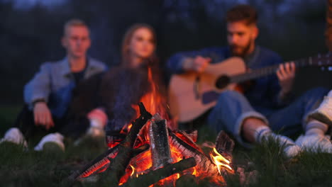 orange flashes of bonfire in the summer evening. company of friends spending good time together sitting by the fire, playing guitar and singing in blur.