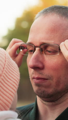 un conmovedor momento al aire libre entre un padre e hija mientras ella ajusta sus gafas mientras lo abraza. el hombre, con una chaqueta verde, la mira con amor mientras ella está cerca