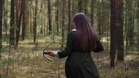 una donna in abito lungo cammina nella foresta con una balestra in mano