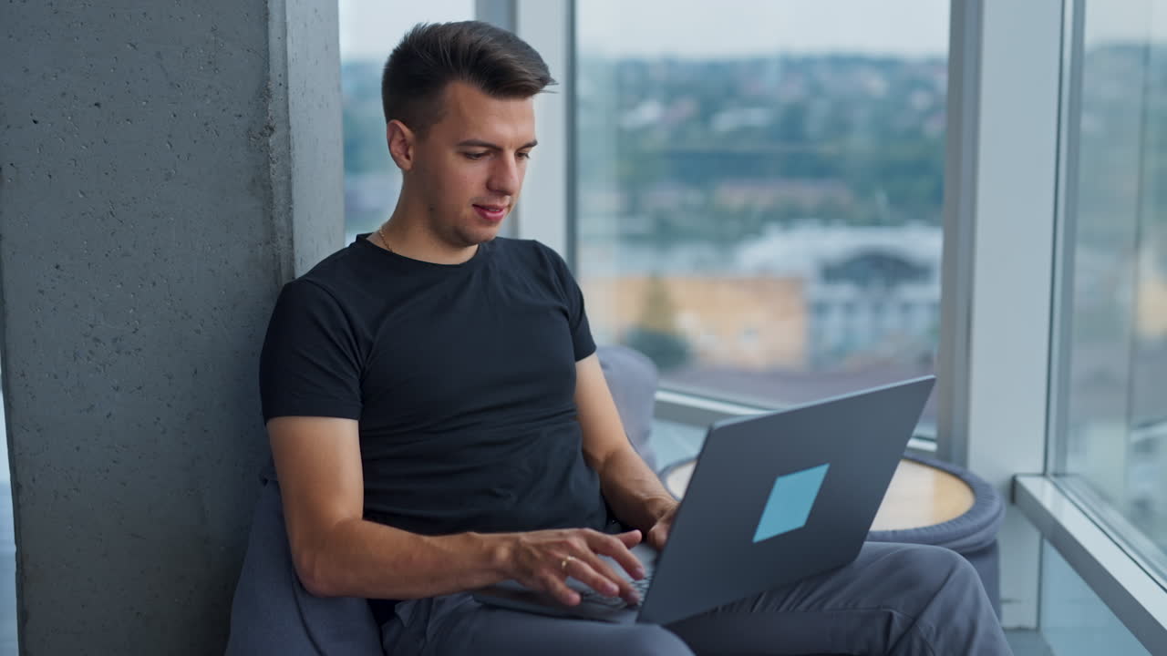 Dark-haired handsome man works on his laptop in a cozy chair. Focused male sits leaned on the concrete pillar in office. Blurred cityscape at backdrop.