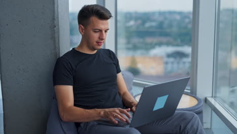 dark-haired handsome man works on his laptop in a cozy chair. focused male sits leaned on the concrete pillar in office. blurred cityscape at backdrop.