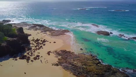 Aerial-tilting-down-view-of-a-wild-sandy-beach-with-rocks,-turquoise-blue-water,-people-and-waves