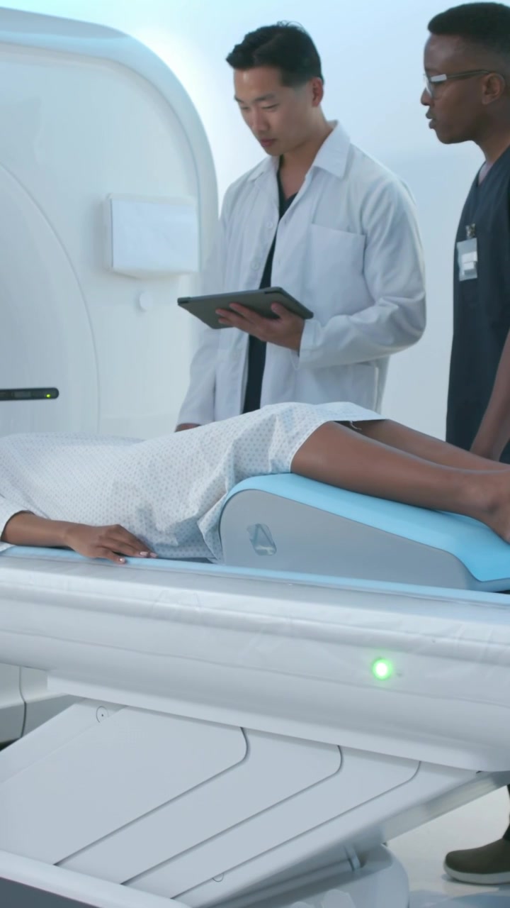 Vertical video: Lying patient in gown on table while doctor reading tablet moving wedge for scanner