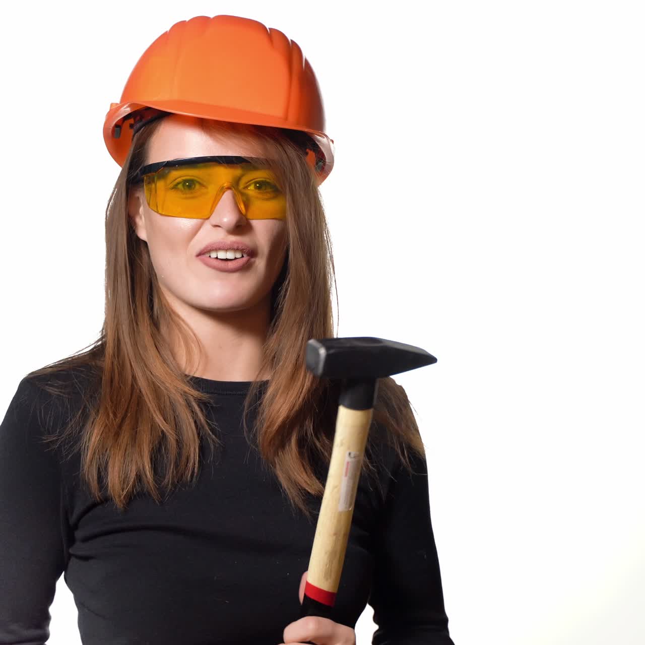 A beautiful woman in orange eyeglasses and helmet is holding a hammer in her hands and hitting herself by it in her hand on a white background in the studio. Engineer