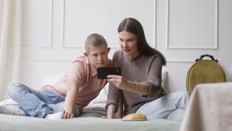 top view of a boy with down syndrome and his mother watching something on smartphone lying on the bed in the bedroom at home