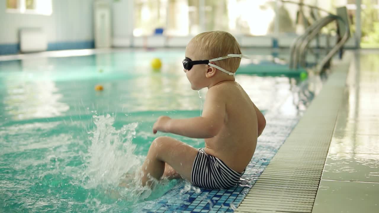 금발 의 작은 소년 이 물 에 발 을  ⁇ 고 수영장 근처 에 앉아 있다. 발 을  ⁇ 고 물 을  ⁇ 고 있다. 측면 전망. 호텔 수영장. 느린 움직임