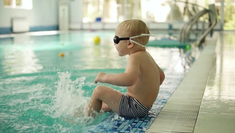 a little boy with blond hair sits by the pool with his feet in the water. dabbles, splashes water with his feet. side view. hotel swimming pool. slow motion