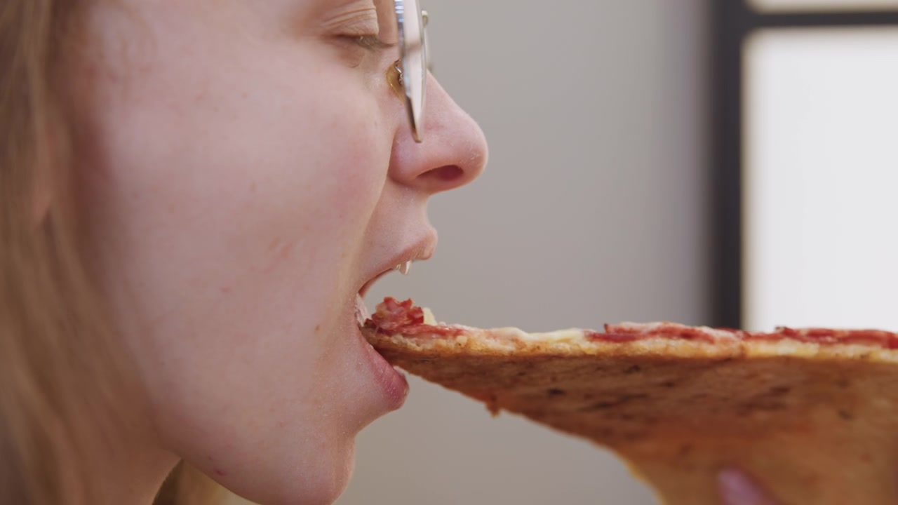 Caucasian woman bites pepperoni slice, glassesclad foodie tasting slice in studio setting, profile closeup on lips and mouth showing subtle smile and satisfied expression, warm indoor lighting