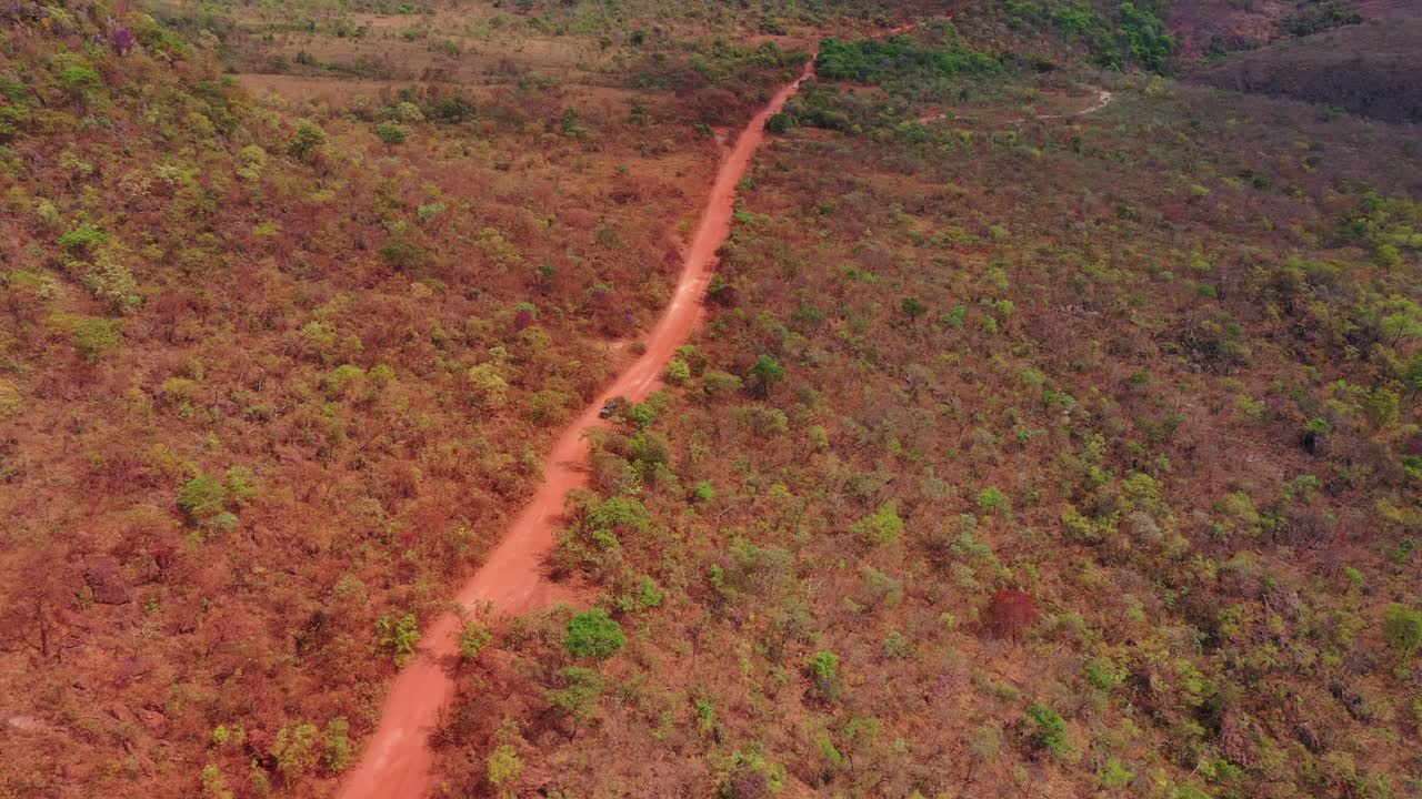 Drone tracks 4x4 penetrating red-soil road through cerrado woodland with rocky ridge escarpment forming dramatic background, red latosoil exposed, dry season in Tocantins central plateau
