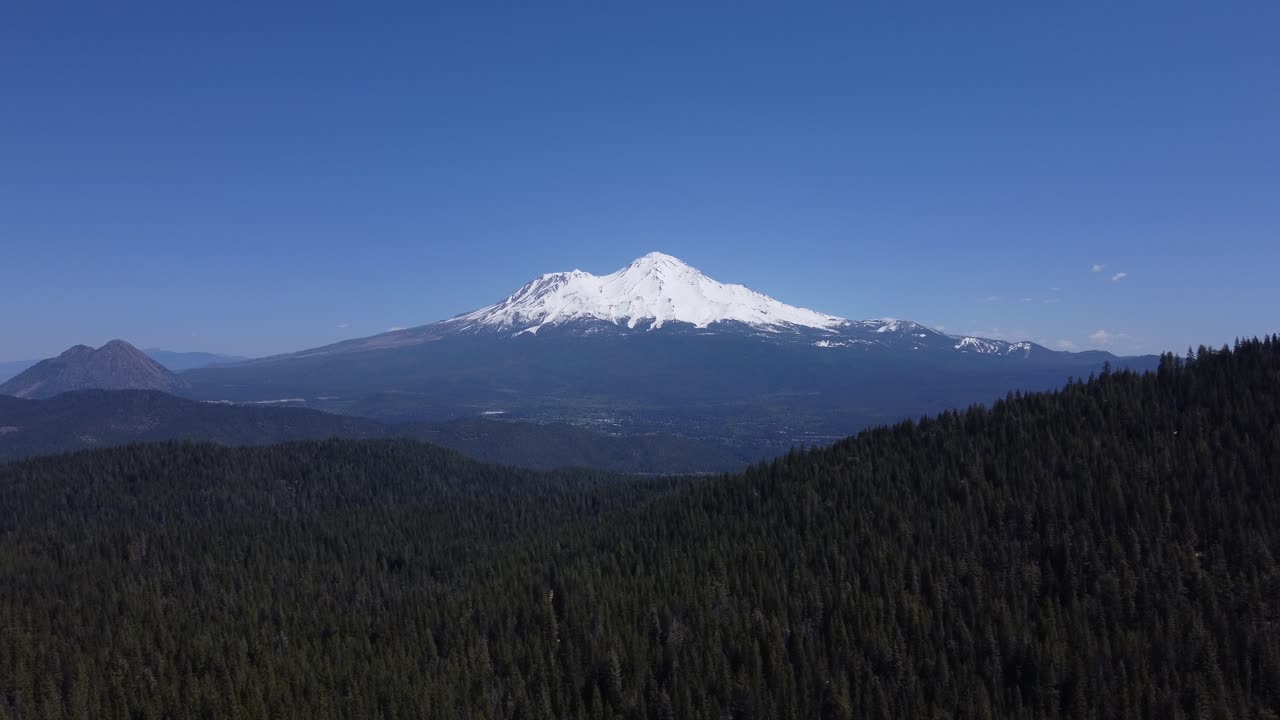 Mount Shasta from the forest in spring in California. Contrast of blue sky, green trees, and white snow glaciers on the peak. Shot in 4K focusing on the mountain towering over the valley.