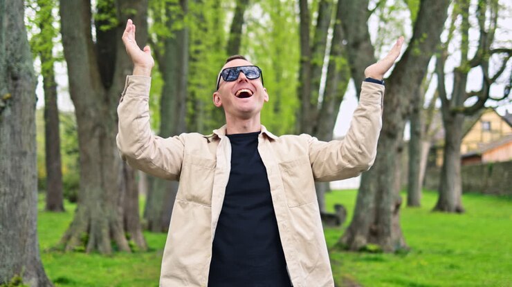 Un uomo con occhiali da sole e una giacca, con un sorriso sul viso, cammina nel parco. Un uomo si toglie gli occhiali da sole e guarda la telecamera mentre cammina all'aperto. Un uomo allegro