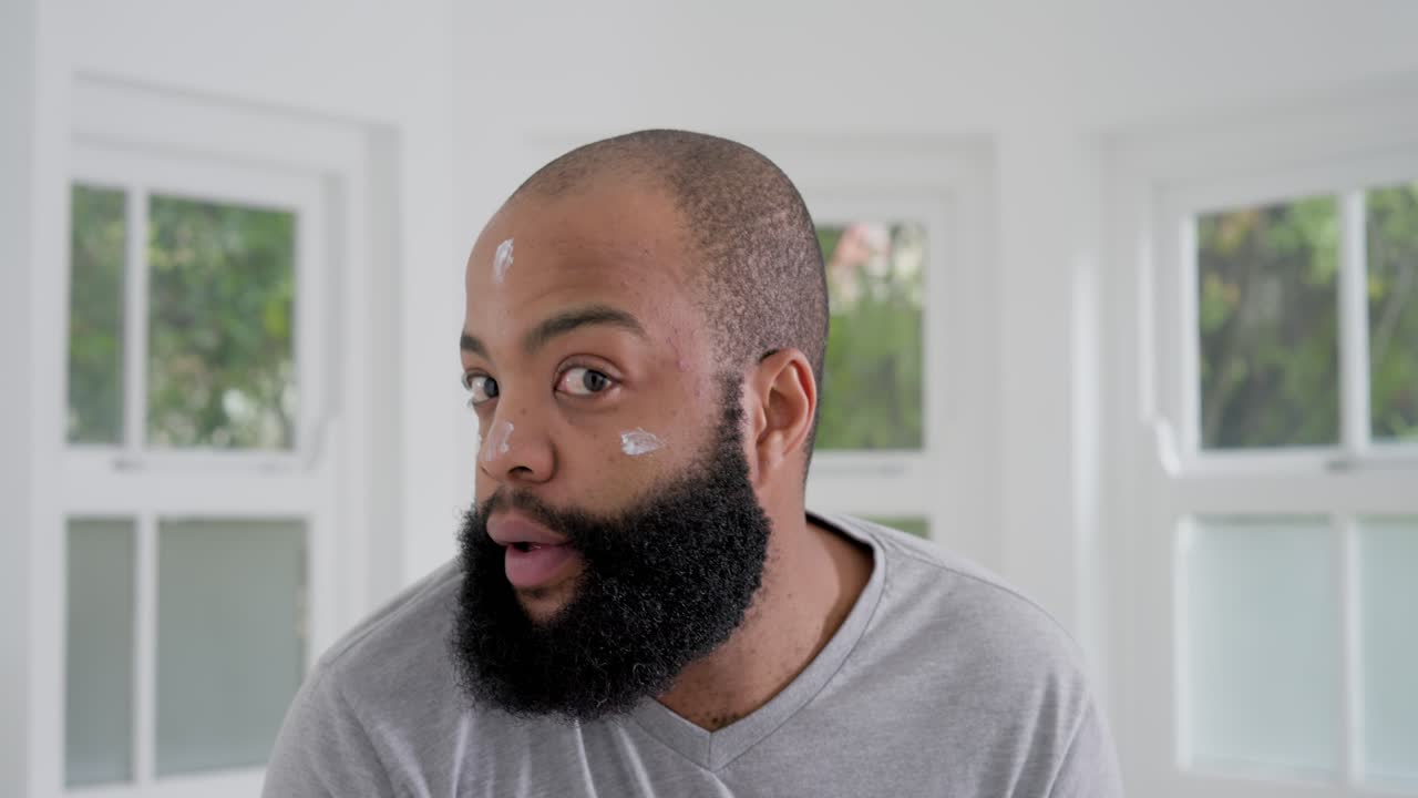 African man in gray shirt at home by windows starting from cheek-cream-dot, dabbing for skincare